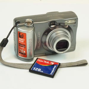 Könnte beinhalten: Eine graue Canon PowerShot A40 Digitalkamera mit silbernem Griff, 3-fach-Zoomobjektiv und Handschlaufe. Die Kamera hat ein rotes Direct Print-Etikett und wird von einer schwarzen SanDisk 128MB CompactFlash-Speicherkarte mit blau-rotem Etikett begleitet.