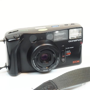 May include: Black Olympus Zoom 35-70mm camera with a lens, flash, and strap. The camera has a zoom lens and flash mode settings. The text "OLYMPUS" and "ZOOM" are visible on the camera body.
