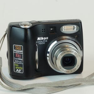 May include: A black Nikon Coolpix 7600 digital camera with a silver lens and a gray wrist strap. The camera has a 7.1 megapixel resolution and a 3x zoom Nikkor ED lens.