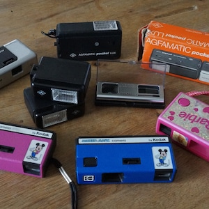 May include: A collection of vintage cameras and flashes, including several Agfamatic Pocket Lux models and Kodak Mickey-Matic cameras. The cameras are in various colors, including black, silver, pink, and blue. One pink camera features a Barbie design.