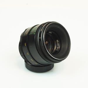 Peut inclure: Un objectif photo noir Helios-44-2 doté d'un élément en verre circulaire, d'une bague de mise au point striée et d'une bague d'ouverture avec des marquages numériques blancs et verts. Le fût affiche des chiffres jaunes et un bord argenté.