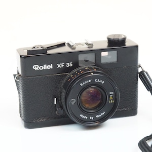 May include: A black Rollei XF 35 camera with a Sonnar 2,3/40 lens. The camera has a black leatherette body, a silver-colored viewfinder, and a black strap. The lens is black with white lettering.