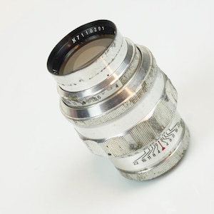 Può includere: Un obiettivo per fotocamera vintage argentato con dettagli neri e segni visibili. L'obiettivo ha una serie di anelli con numeri e un bordo nero con la scritta "N7110201". L'obiettivo è posto su uno sfondo bianco.