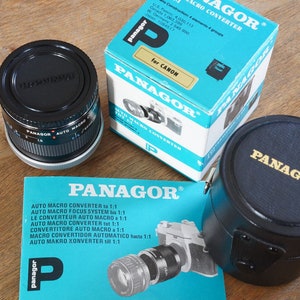 Puede incluir: Un objetivo de cámara negro con un anillo plateado y una tapa de objetivo negra. El objetivo está etiquetado como "Panagor Auto Macro 1:1". El objetivo está en una funda de cuero negro con una etiqueta dorada "Panagor". El objetivo también está en una caja azul con el texto "Panagor Auto Macro Converter for Canon" impreso en ella.