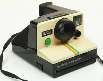 May include: A black and cream Polaroid 1000 Land Camera featuring a prominent black lens, a bright green shutter button, and a signature rainbow stripe graphic on the front panel. The device includes a black carrying strap attached to the side.