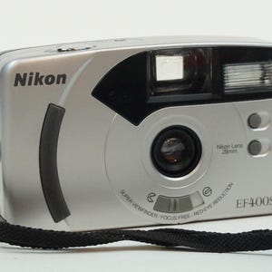 Puede incluir: Una cámara de fotos compacta Nikon EF400SV de color plateado con una lente negra y una correa negra. La cámara tiene un flash incorporado y una función de reducción de ojos rojos.