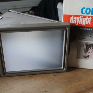 May include: A gray Copal daylight screen DS-2 for movies, slides, or filmstrips. The screen has a rectangular viewing area and a box with the product name and a picture of a child on a horse. The screen is on a wooden surface.