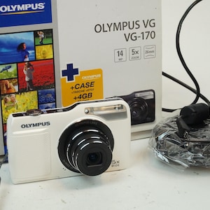 May include: White Olympus VG-170 digital camera with a black lens and a flash. The camera is next to its box, which displays the camera model and features. A charger and cable are also included.