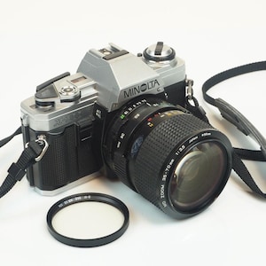 Puede incluir: Una cámara de película Minolta X-300 plateada y negra con un objetivo zoom de 35-70 mm. La lente tiene un agarre negro texturizado. Un filtro de lente negro se encuentra frente a la cámara. La correa de la cámara es negra.