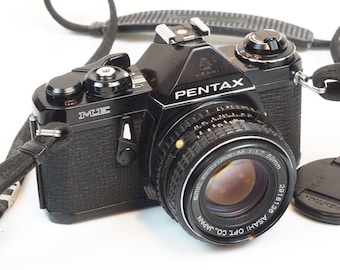 Peut inclure: Appareil photo argentique Pentax ME noir avec un objectif de 50 mm. L'appareil photo est équipé d'une sangle noire et d'un bouchon d'objectif. L'objectif porte l'inscription "SMC PENTAX-M 1:1.7 50mm" et "ASAHI OPT. CO., JAPAN".