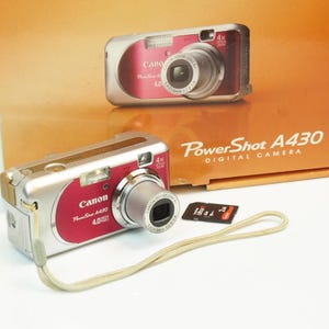 May include: A silver and red Canon PowerShot A430 digital camera with a 4x zoom lens. The camera has a wrist strap and a memory card. The box behind the camera reads "PowerShot A430 DIGITAL CAMERA".
