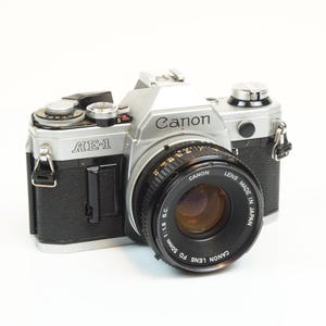 Puede incluir: Una cámara Canon AE-1 con cuerpo negro y plateado y una lente Canon FD 50mm 1:1.8 S.C. La cámara tiene el texto "Canon" y "AE-1" impreso. La lente es negra con texto blanco y está hecha en Japón.