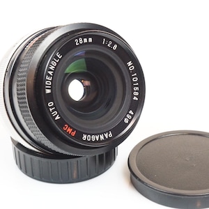 May include: A black and silver Panagor PMC 28mm wide-angle auto camera lens with a black lens cap. The lens has the following text: "28mm 1:2.8", "WIDEANGLE AUTO", and "NO.101584". The lens cap is round.