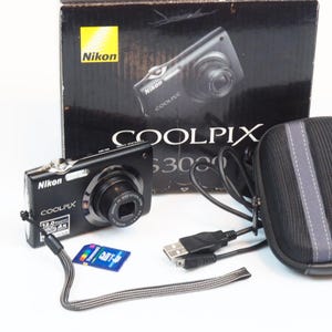 Op de afbeelding: Een zwarte Nikon Coolpix digitale camera met een 12,0 megapixel lens, een polsbandje, een blauwe geheugenkaart, een zwarte USB-kabel en een zwarte etui met ritssluiting, weergegeven voor de originele Nikon Coolpix S3000 productdoos.