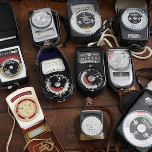 May include: A collection of vintage light meters, including a Sekonic, a Gossen, a  Lumu, a  Wiltz, a  Polysix, and a  Sonoflex. The light meters are all in black, silver, and white cases with various dials and buttons.