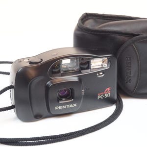 Puede incluir: Cámara autofocus Pentax PC-50 negra con correa negra y funda de cuero negra. La cámara tiene el logotipo de Pentax y "AF PC-50" impreso en la parte delantera. La cámara está sobre un fondo blanco.
