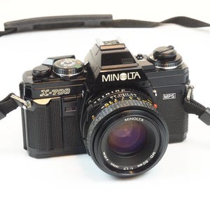 Könnte beinhalten: Eine schwarze Minolta X-700 35mm Filmkamera mit einem angebrachten Minolta MD 50mm 1:1.7 Objektiv. Das Kameragehäuse zeigt das Minolta-Logo, den X-700 Modellschriftzug und ein MPS-Abzeichen. Ein schwarzer Trageriemen ist an den Ösen befestigt.