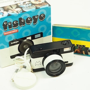 May include: A black and white Fisheye Lomography camera with a white lens and a matching lens cap. The camera is next to its original box, which has a blue background and the word "fisheye" in white bubble letters.