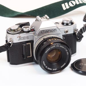 Puede incluir: Una taza de cerámica blanca con asa negra y el texto "But First, Coffee" en negrita negra. La taza se encuentra sobre un fondo borroso, sugiriendo un ambiente acogedor.