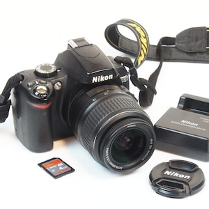 Può includere: Una fotocamera reflex digitale Nikon D60 nera con obiettivo AF-S DX Nikkor 18-55mm collegato, una tracolla Nikon nera, un caricabatterie rapido Nikon MH-23 nero, un copriobiettivo nero con logo Nikon e una scheda di memoria SD SanDisk Ultra 4GB.