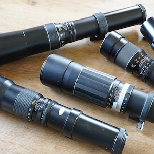 Puede incluir: Cinco objetivos de cámara negros de diferentes tamaños y longitudes, algunos con marcas y números, sobre una superficie de madera.