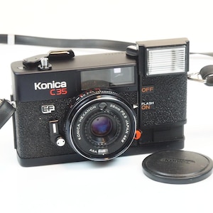 May include: Black Konica C35 camera with a 38mm lens and a flash. The camera has the text "Konica C35" in white and red. A lens cap is next to the camera. The camera strap is visible.
