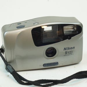 May include: A silver Nikon BF100 film camera with a black wrist strap. The camera features a lens, flash, and buttons. The text "Nikon BF100" is printed on the front of the camera.