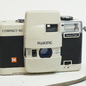 May include: A vintage Haking Compact-SC camera with a cream-coloured body and black leatherette accents. The camera features the text "Haking" and "Compact-SC", a lens with f/3.5-38mm, and a built-in flash. Includes a grey wrist strap.