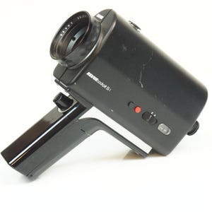 Peut inclure: Une caméra de cinéma vintage noire Revue cockpit S1. La caméra possède un objectif, une poignée et plusieurs boutons et interrupteurs. Le texte "REVUE cockpit S1" est imprimé sur le côté. L'objectif porte l'inscription "REVUE OBJECTIVE".