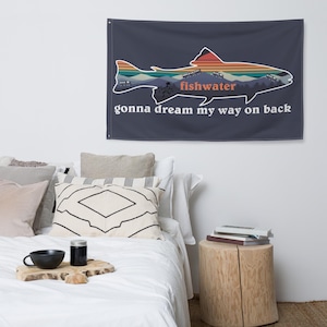 Op de afbeelding: Een marineblauwe wandvlag met een visvormig ontwerp met een berg- en zonsondergangscène. Het woord "fishwater" is oranje en de zin "gonna dream my way on back" is wit.