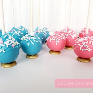 May include: A close-up of cake pops decorated with sprinkles. The cake pops are arranged in two rows, with the top row being blue and the bottom row being pink. The cake pops are on sticks and have a gold base.