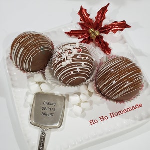 May include: Three chocolate hot cocoa bombs decorated with white chocolate and sprinkles on a white plate with a silver spoon that says "Baking Spirits Bright" and white marshmallows. The plate also has the text "Ho Ho Homemade" in red.