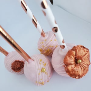 May include: Three pink cake pops with gold sprinkles and a rose-gold flower on one. The cake pops are on sticks with white and gold striped paper straws.