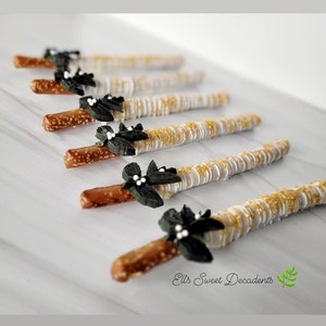 May include: A close-up of six pretzel rods dipped in white chocolate and covered in gold sprinkles. Each pretzel rod is decorated with a black and white leaf-shaped embellishment.