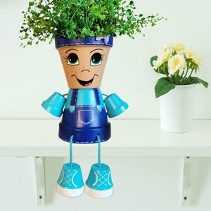 May include: A blue and turquoise painted flower pot with a smiling face, wearing overalls and blue sneakers. The pot is sitting on a white shelf and has a green plant growing out of the top.