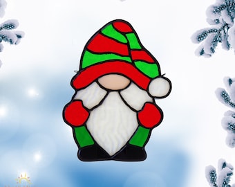 Op de afbeelding: Een ornament van gekleurd glas met een dwerg met een rood-groen gestreepte hoed, witte baard en rode wanten. De dwerg is tegen een besneeuwde achtergrond.