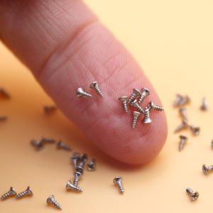 May include: Close-up of small silver screws scattered on a light orange surface, with some resting on a fingertip. The screws have a slotted head and a pointed end. The image highlights the small size and quantity of the hardware.