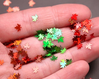 May include: A hand holding a variety of colorful glitter maple leaves. The leaves are in shades of red, orange, green, and yellow.