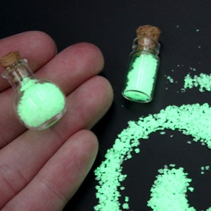 May include: Two small glass bottles with cork stoppers filled with glowing green powder. A small pile of the powder is scattered on a black surface.