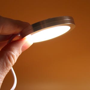 May include: A small, round, silver-colored LED light is held in a hand. The light is emitting a bright, white glow. A white cord is attached to the light. The background is a warm, orange color.