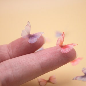 May include: A fingertip holds two delicate butterfly-shaped embellishments. One butterfly is pink and the other is purple. The butterflies are made of a translucent material and have a delicate, feminine appearance.