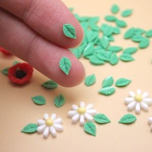 May include: A collection of miniature floral embellishments, including white daisies with yellow centers, red poppies, and green leaves. The leaves are held between fingers, showcasing the detail of the craft supplies. The items are scattered on a light tan surface.