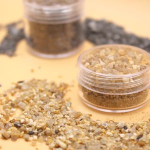 May include: Two small, clear plastic jars filled with small, light-colored gravel. The jars sit on a light orange surface, with scattered gravel around them. A third jar with darker gravel is in the background.