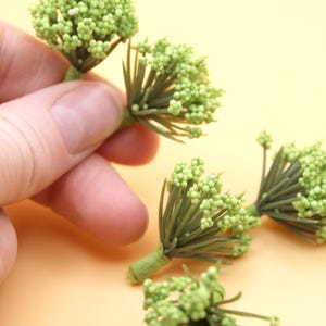 May include: Close-up of several small, light green artificial flower stems. Each stem features a cluster of tiny, round buds and thin, green leaves. The stems are held by a hand against a light orange background.