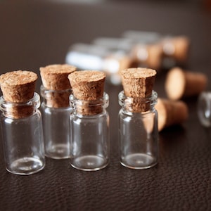May include: Small, clear glass bottles with cork stoppers. Several bottles are upright, while others are scattered on a dark brown surface. The corks are a light brown color, and the bottles appear empty.