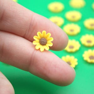 May include: A miniature yellow sunflower with a brown center is held between fingers. The flower has bright yellow petals and a detailed center. Several similar flowers are scattered on a green surface, suggesting a craft or decorative theme.