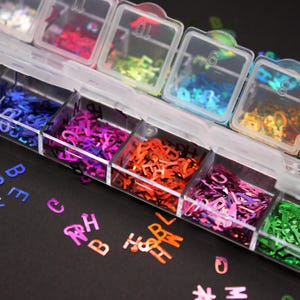 May include: A clear plastic organizer filled with colorful letter-shaped sequins. The sequins are in various colors, including red, orange, purple, blue, and green. The organizer has numbered compartments, and some sequins are scattered on a black surface.