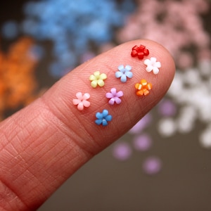 May include: A fingertip displays a collection of colorful flower-shaped beads. The beads are in shades of red, orange, yellow, green, blue, and purple.