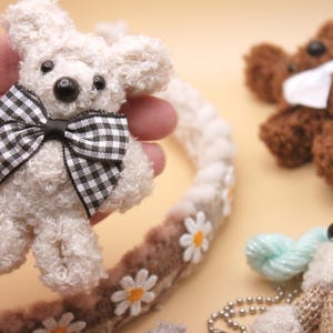May include: A white teddy bear with a black and white checkered bow tie. The bear is sitting in a hand and is surrounded by other small stuffed animals.