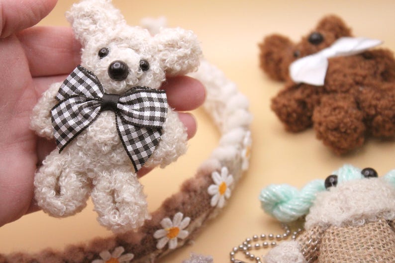 May include: A white teddy bear with a black and white checkered bow tie. The bear is sitting in a hand and is surrounded by other small stuffed animals.